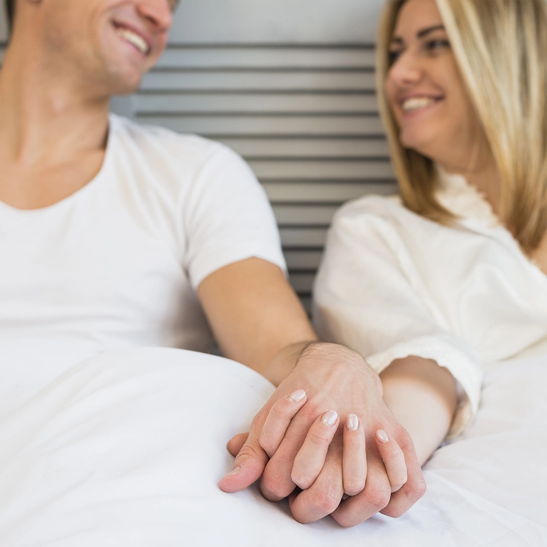 A Couple Holding Hands In Their Bed And Smiling At Each Other
